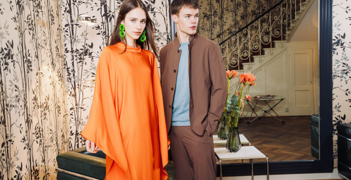 Model in fließendem orangefarbenem Kaftan mit Statement-Ohrringen neben Model in braunem Anzug im stilvollen Interieur – FESCH Editorial „Urbane Leichtigkeit“