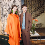Model in fließendem orangefarbenem Kaftan mit Statement-Ohrringen neben Model in braunem Anzug im stilvollen Interieur – FESCH Editorial „Urbane Leichtigkeit“