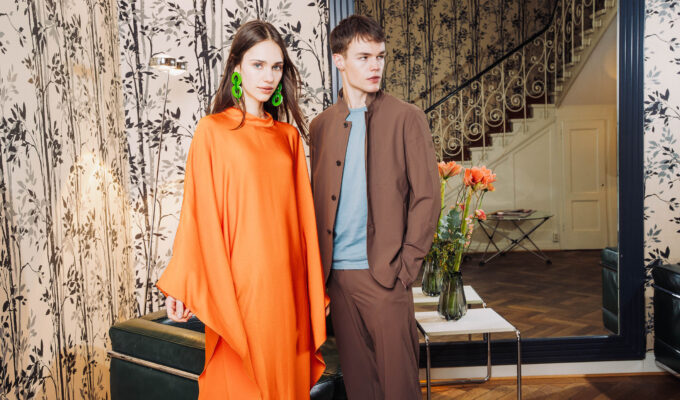Model in fließendem orangefarbenem Kaftan mit Statement-Ohrringen neben Model in braunem Anzug im stilvollen Interieur – FESCH Editorial „Urbane Leichtigkeit“