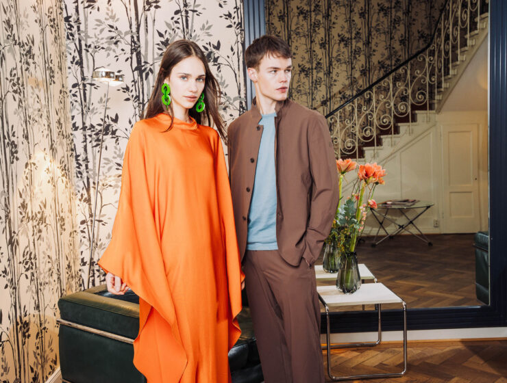 Model in fließendem orangefarbenem Kaftan mit Statement-Ohrringen neben Model in braunem Anzug im stilvollen Interieur – FESCH Editorial „Urbane Leichtigkeit“