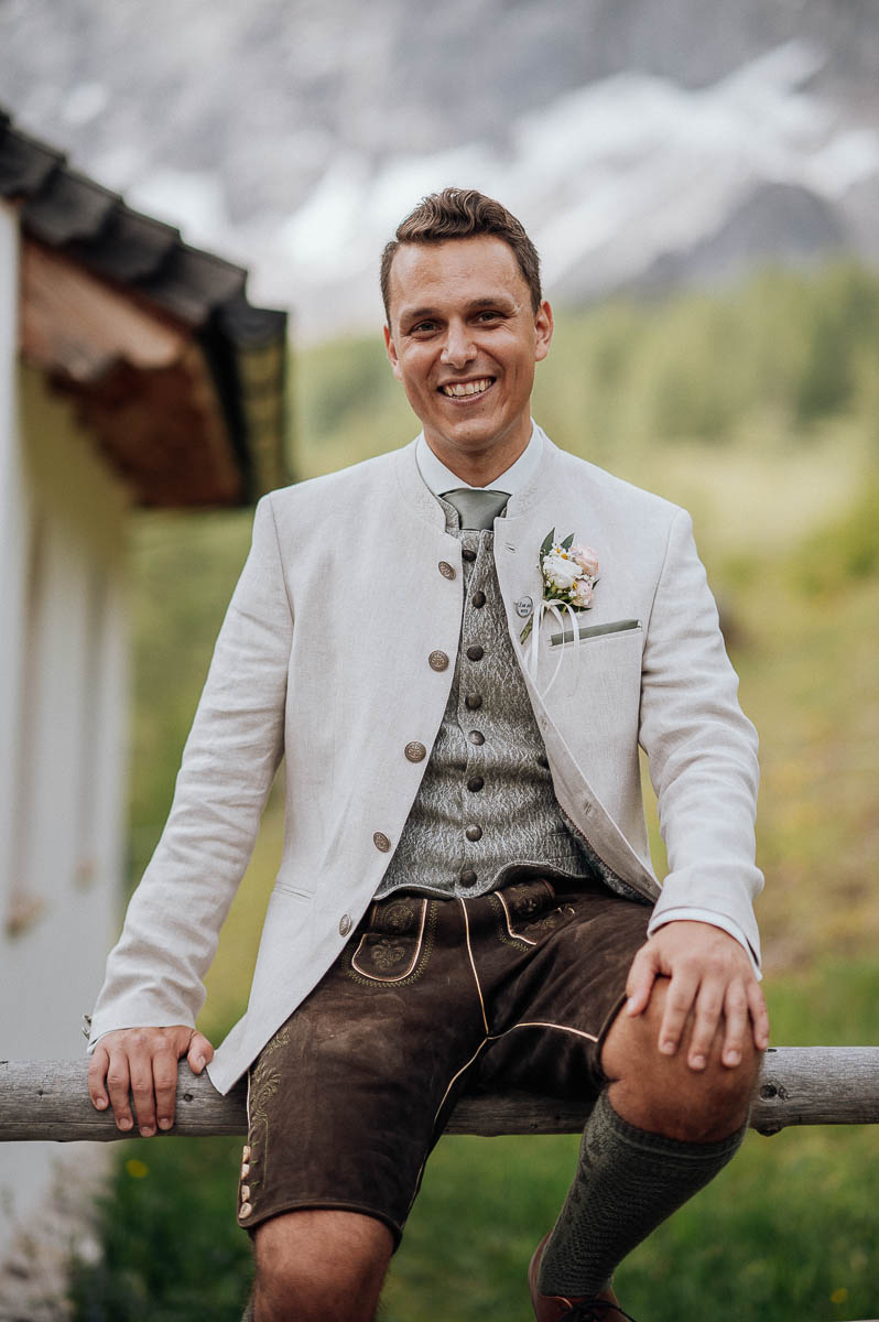 Bräutigam sitzt auf einem Holzzaun vor alpiner Bergkulisse