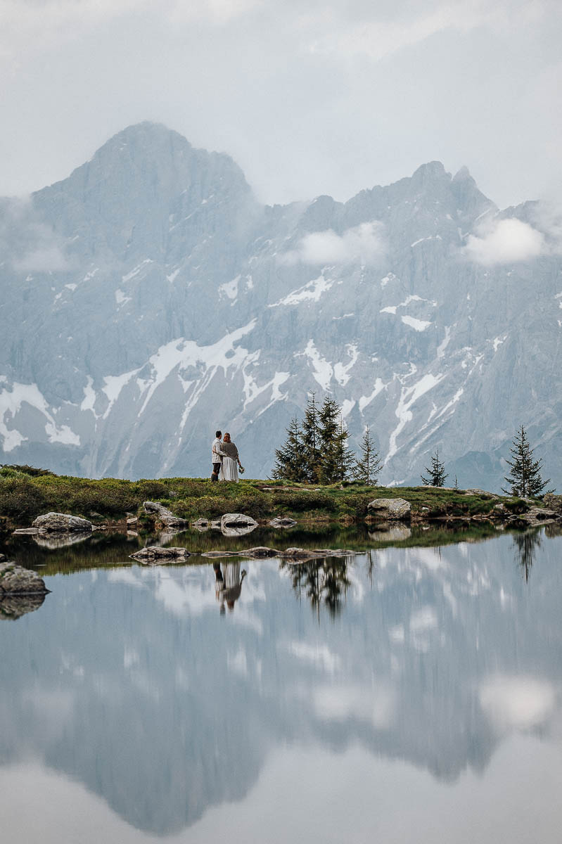 Brautpaar steht an einem Bergsee mit Spiegelung der Berge im Wasser