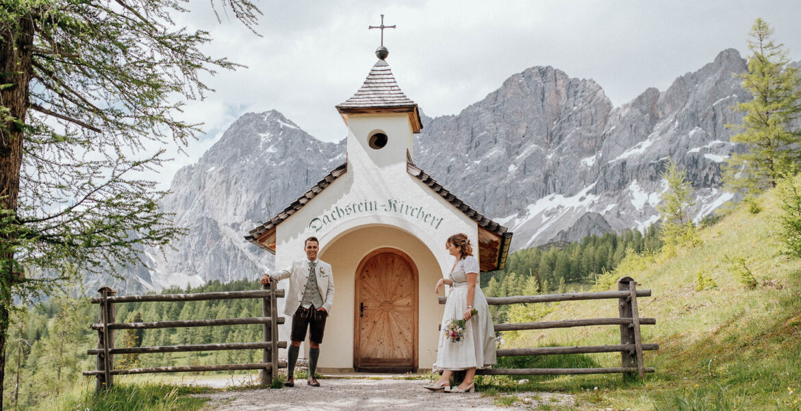Brautpaar in Tracht steht vor der Dachstein Kapelle mit Bergkulisse im Hintergrund