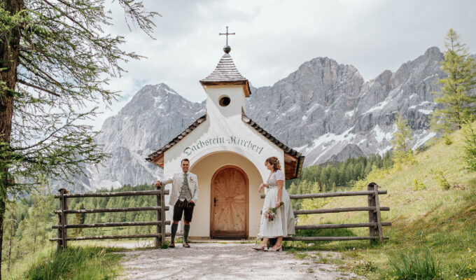 Brautpaar in Tracht steht vor der Dachstein Kapelle mit Bergkulisse im Hintergrund