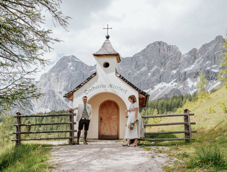 Brautpaar in Tracht steht vor der Dachstein Kapelle mit Bergkulisse im Hintergrund
