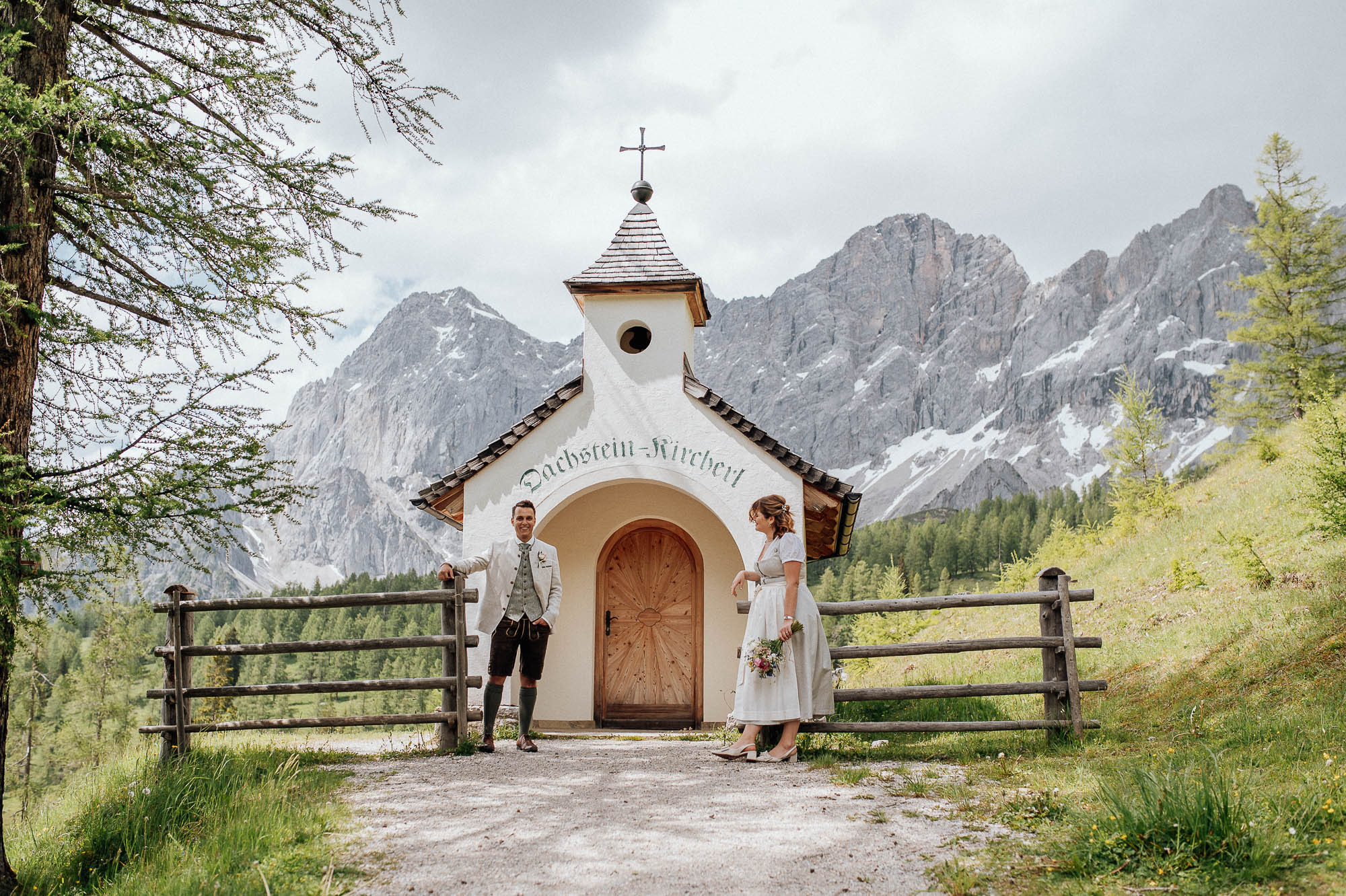 Brautpaar in Tracht steht vor der Dachstein Kapelle mit Bergkulisse im Hintergrund