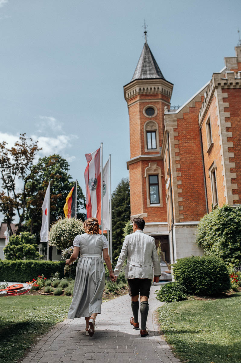 Brautpaar in Tracht läuft Hand in Hand zum Standesamt vor einem Schloss in Schladming