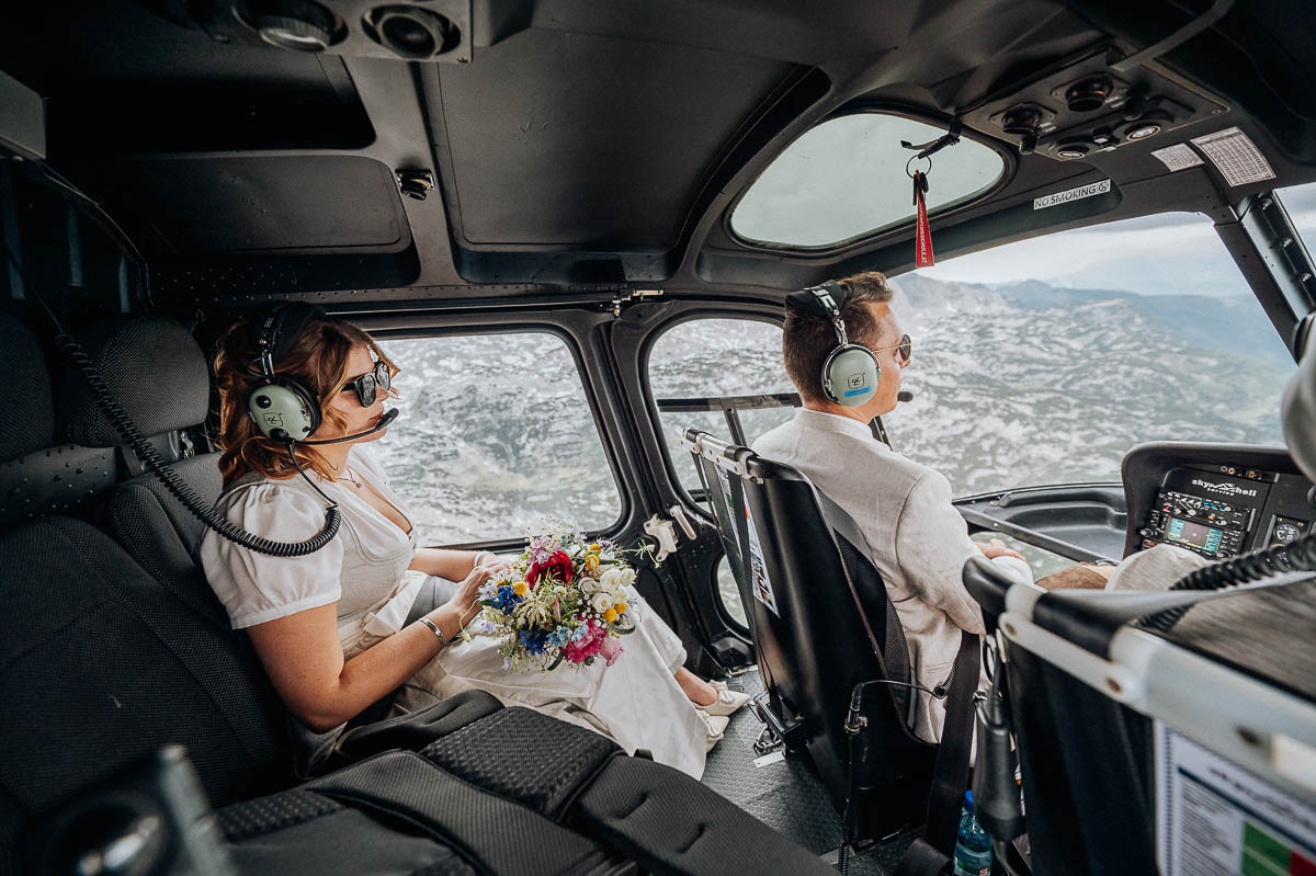 Brautpaar sitzt im Helikopter mit Kopfhörern und Blick auf die Alpen