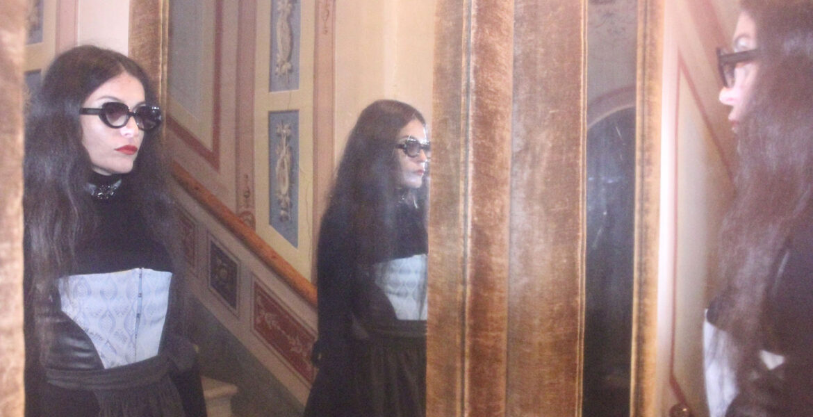 Frau mit Sonnenbrille trägt ein Korsett von MARIAHAUSE und steht vor einem Spiegel in einem historischen Interieur