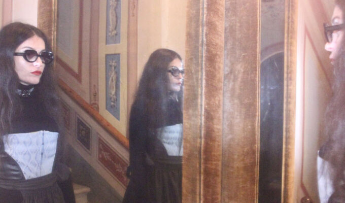 Frau mit Sonnenbrille trägt ein Korsett von MARIAHAUSE und steht vor einem Spiegel in einem historischen Interieur