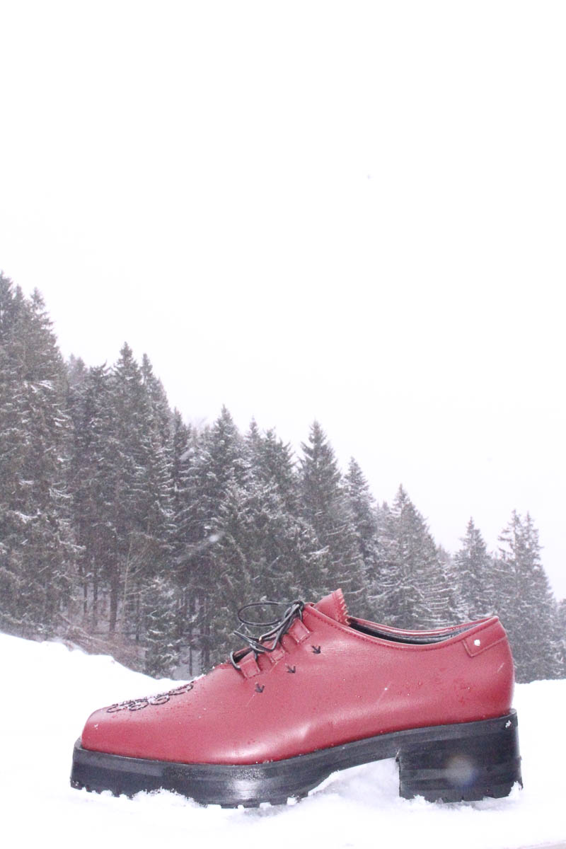 Roter handgefertigter Lederschuh von MARIAHAUSE steht im Schnee vor Waldkulisse