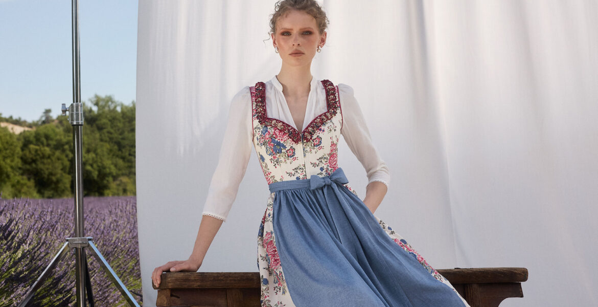 Frau im floralen Dirndl mit blauer Schürze vor hellem Hintergrund und Lavendelfeld – Muttertag 2026 Mode Inspiration