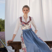 Frau im floralen Dirndl mit blauer Schürze vor hellem Hintergrund und Lavendelfeld – Muttertag 2026 Mode Inspiration