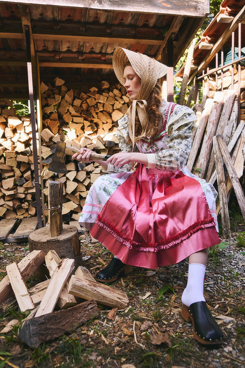 Cottagecore auf dem Land Model im rosa Dirndl beim Holzhacken vor rustikalem Holzlager