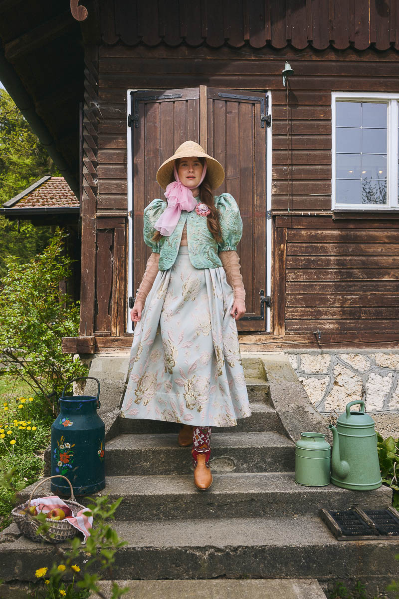 Cottagecore-Tracht am Holzhaus Model in romantischem Dirndl vor traditionellem Holzhaus im Cottagecore-Stil