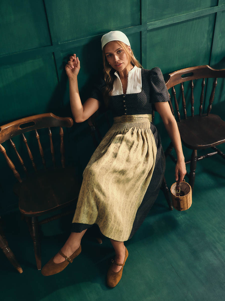 Dirndl Laura und Bluse Josi von Gottseidank Frau im dunklen Dirndl mit heller Schürze und weißer Haube zwischen historischen Holzstühlen