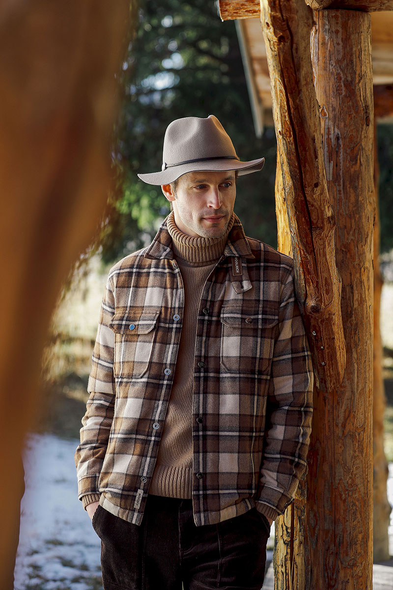 Mann mit braun-beige karierter Hemdjacke, braunem Rollkragenpullover, dunkler Cordhose und hellem Hut vor einer Holzhütte