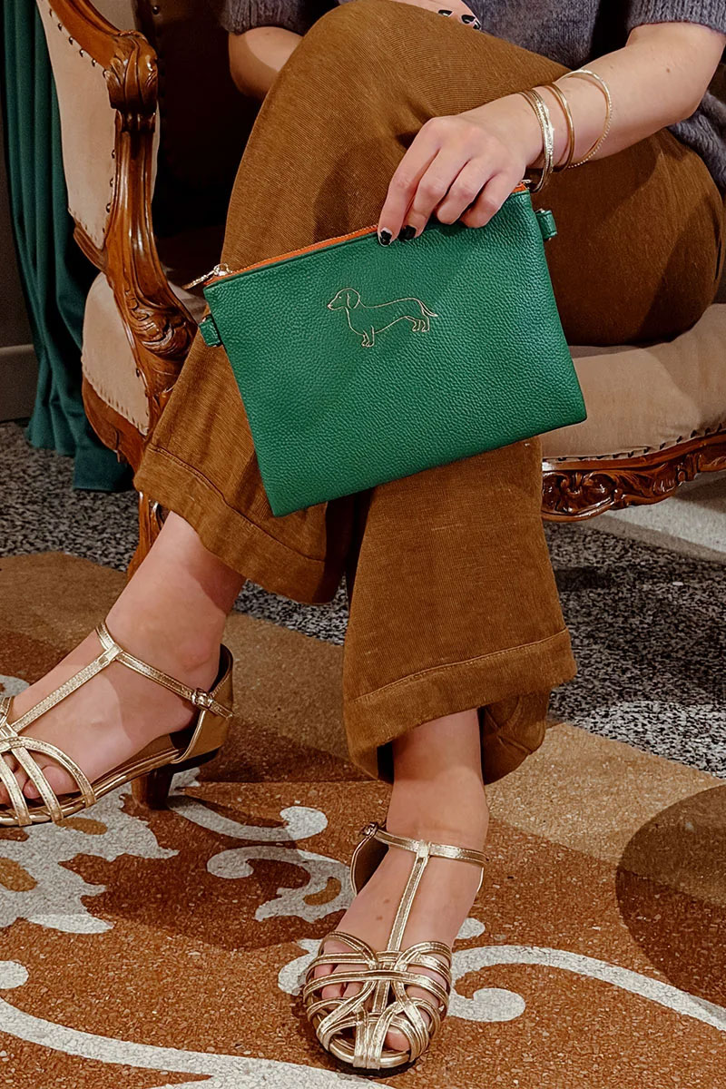 Grüne Pouch von Mimi’s Glow mit goldfarbener Dackelstickerei, kombiniert mit brauner Hose und goldenen Sandalen.